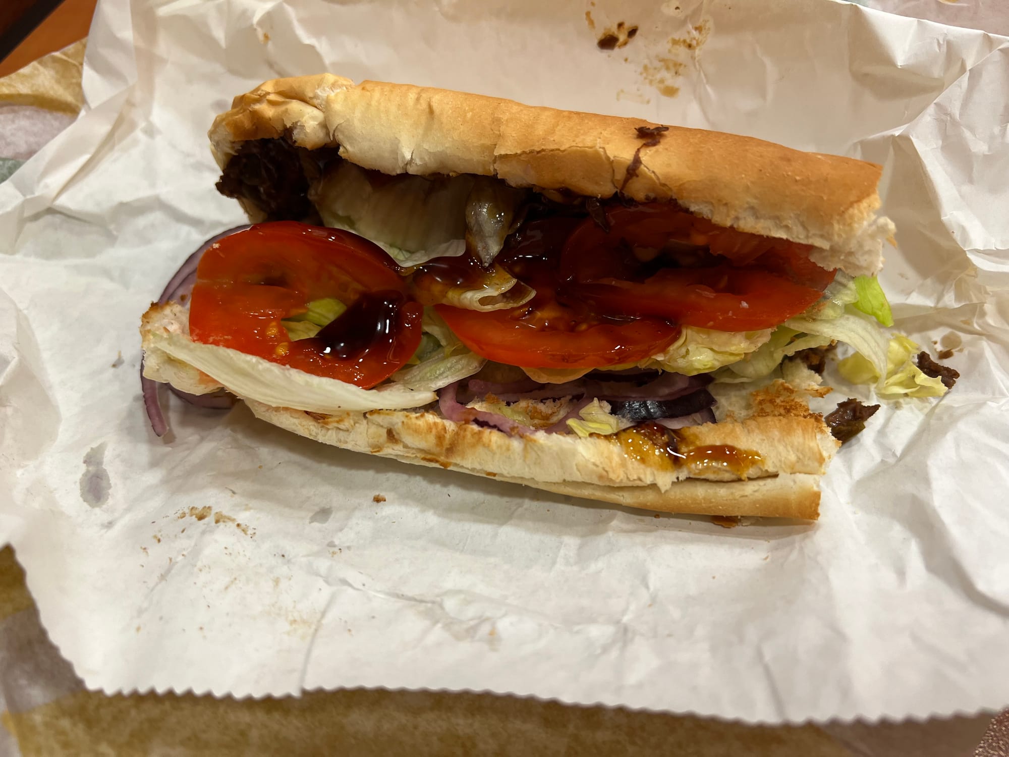 An unappetizing looking Subway sandwich. The bread is all fucked up, theres a brown sauce splattered all over it, and you can't even see the faux meat and cheese.
