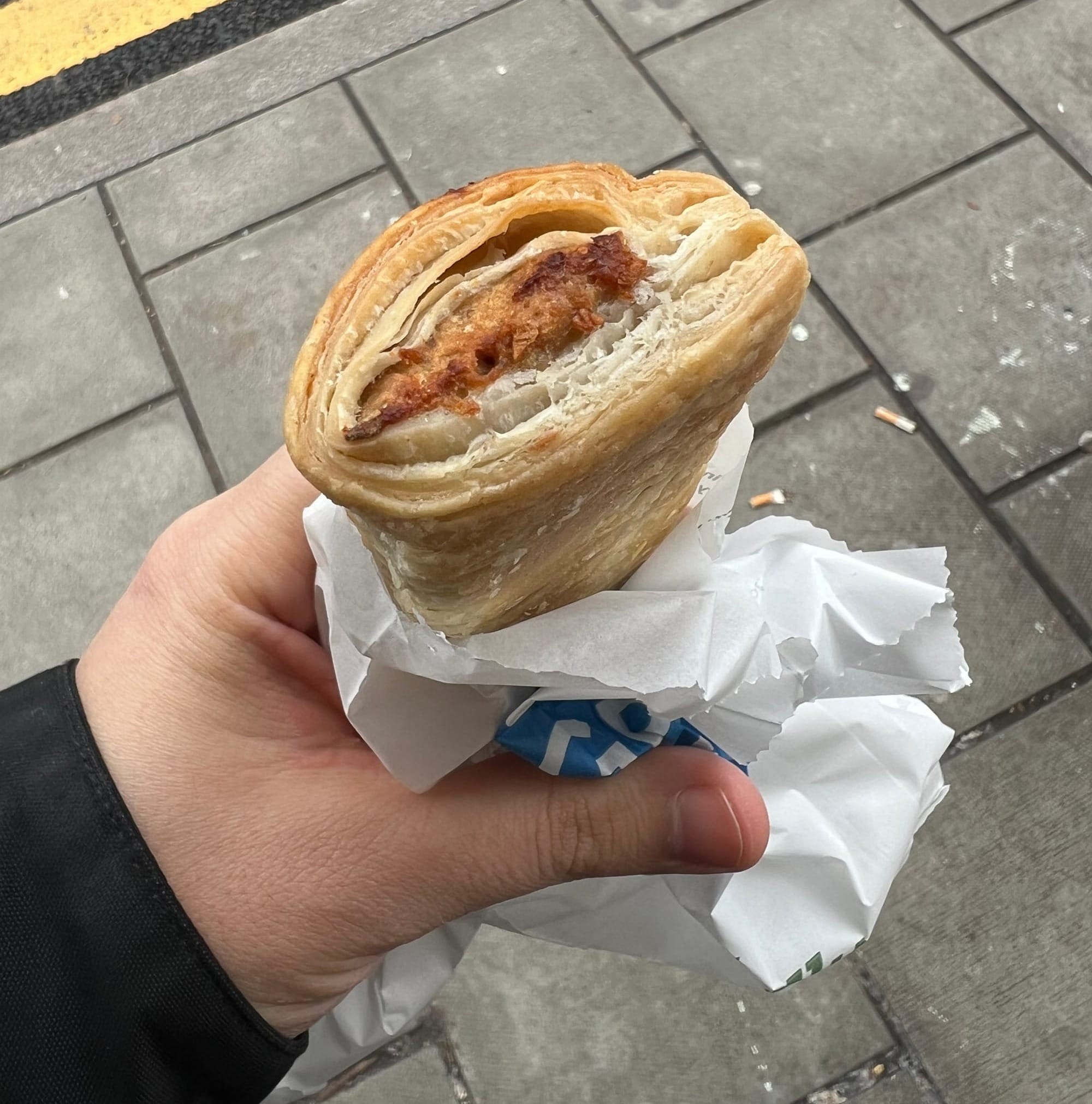 A hand holding a vegan sausage roll wrapped, still in its little wrapper from the store, outside on the sidewalk.