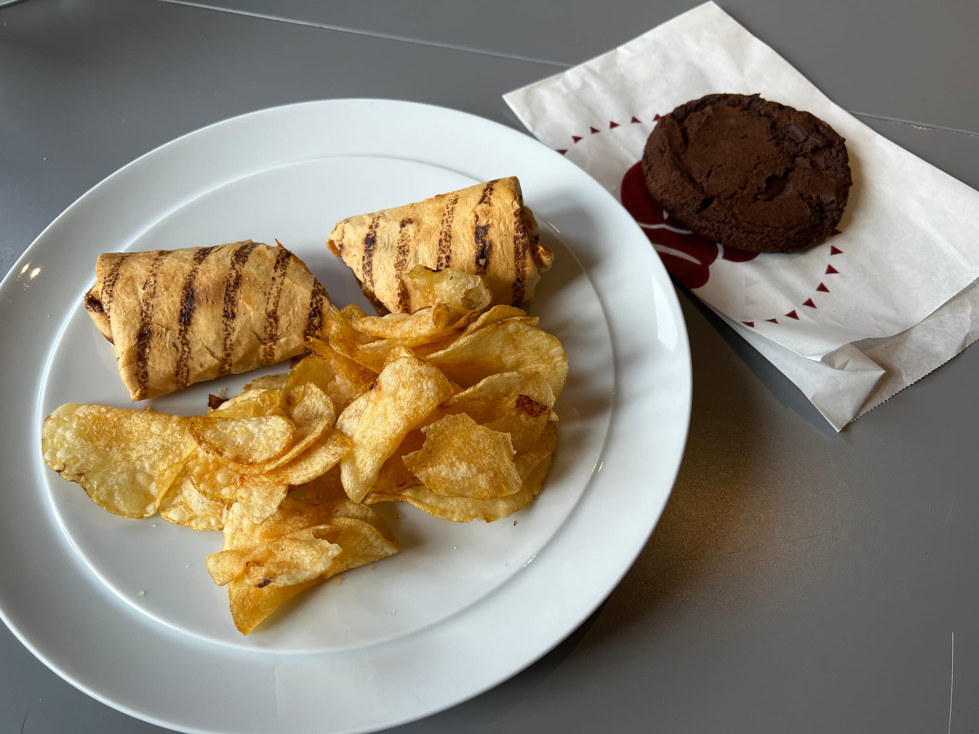A wrap with grill marks on a plate with some crisps. A chocolate cookie on a Costa to-go wrapper sits next to the plate.