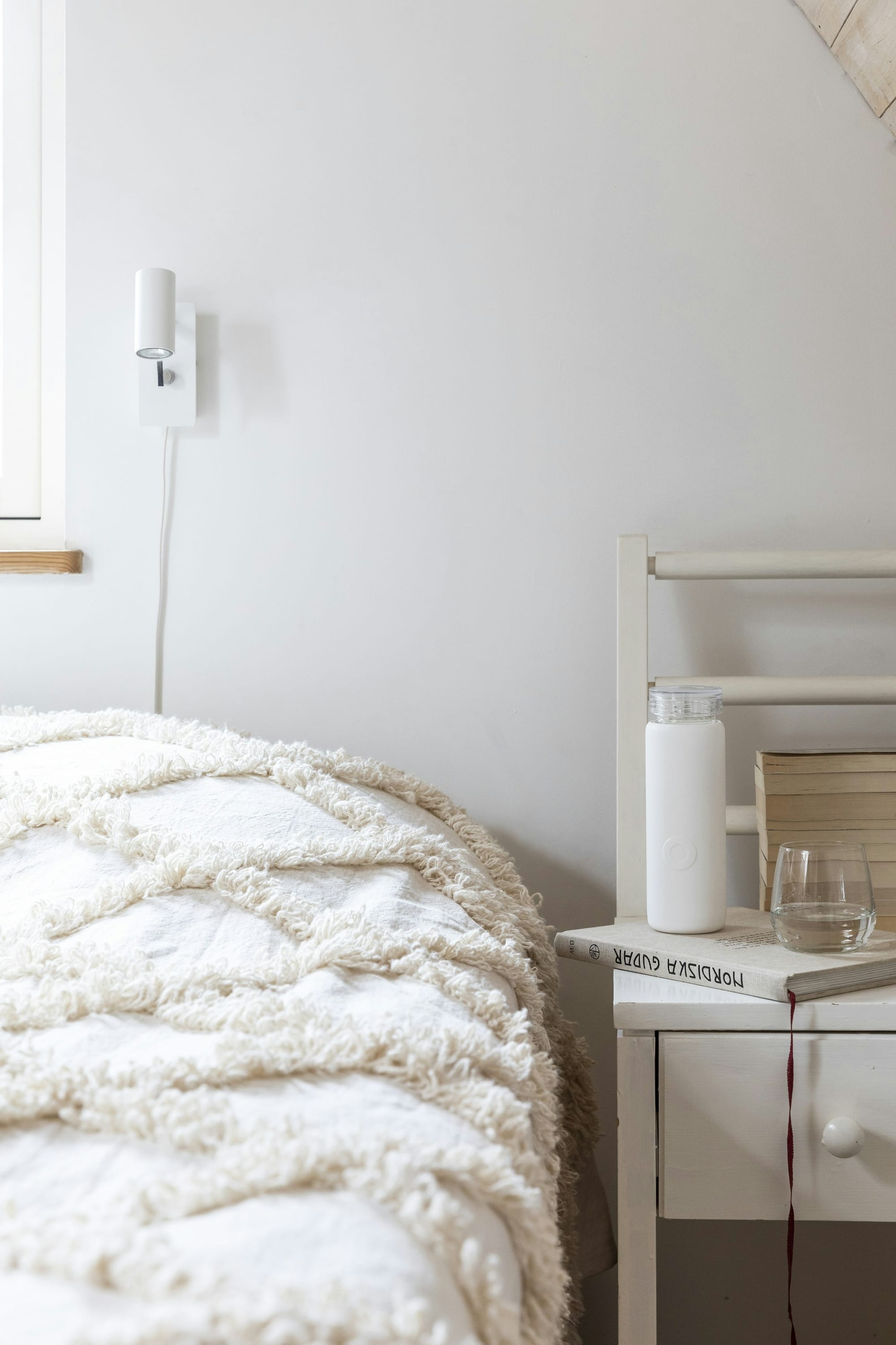 A white water bottle sides on a bedside table next to a bed with some white linens and blankets.