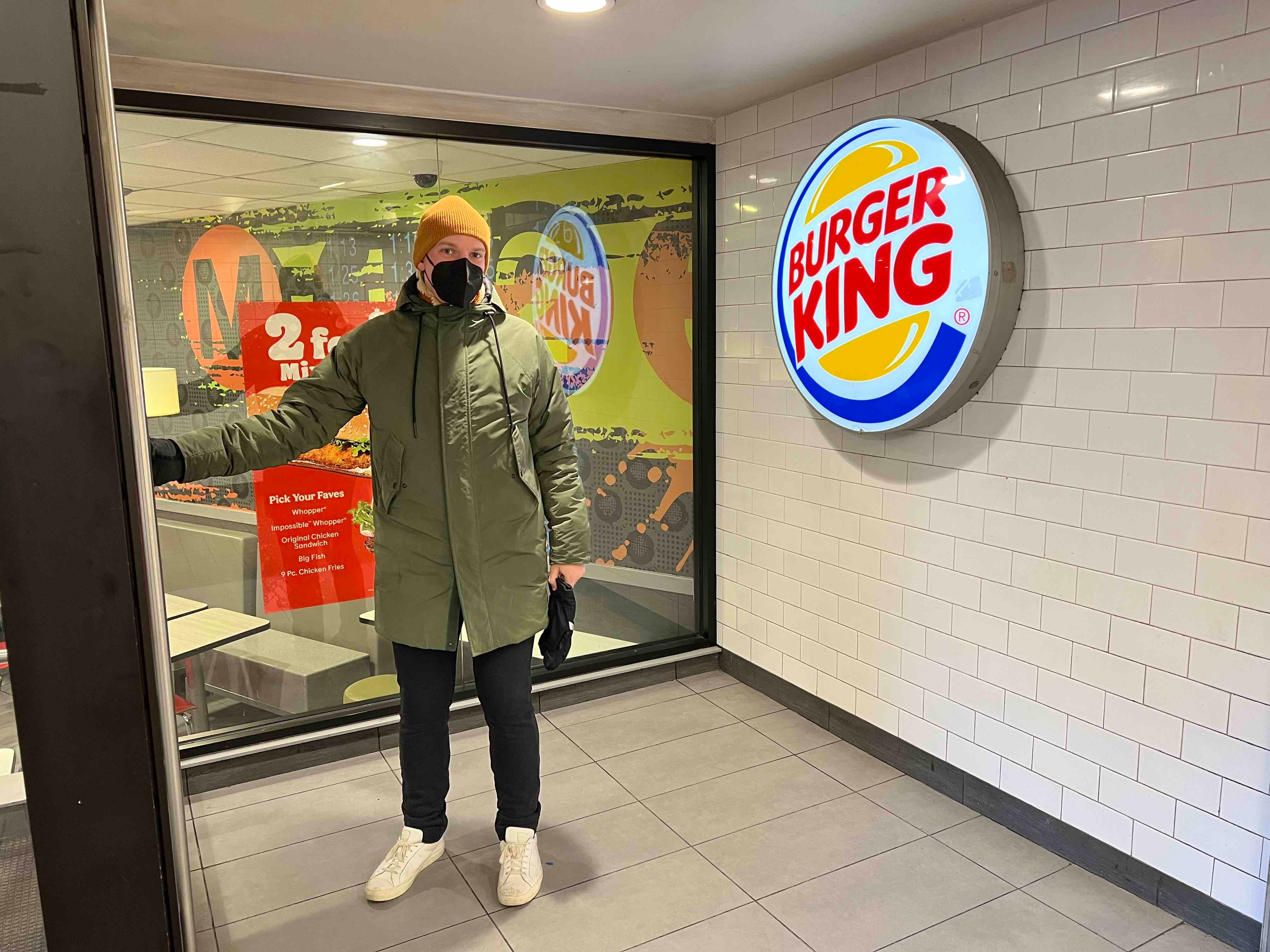 A man in winter clothing stands in front of a Burger King entrance inside of a subway station.