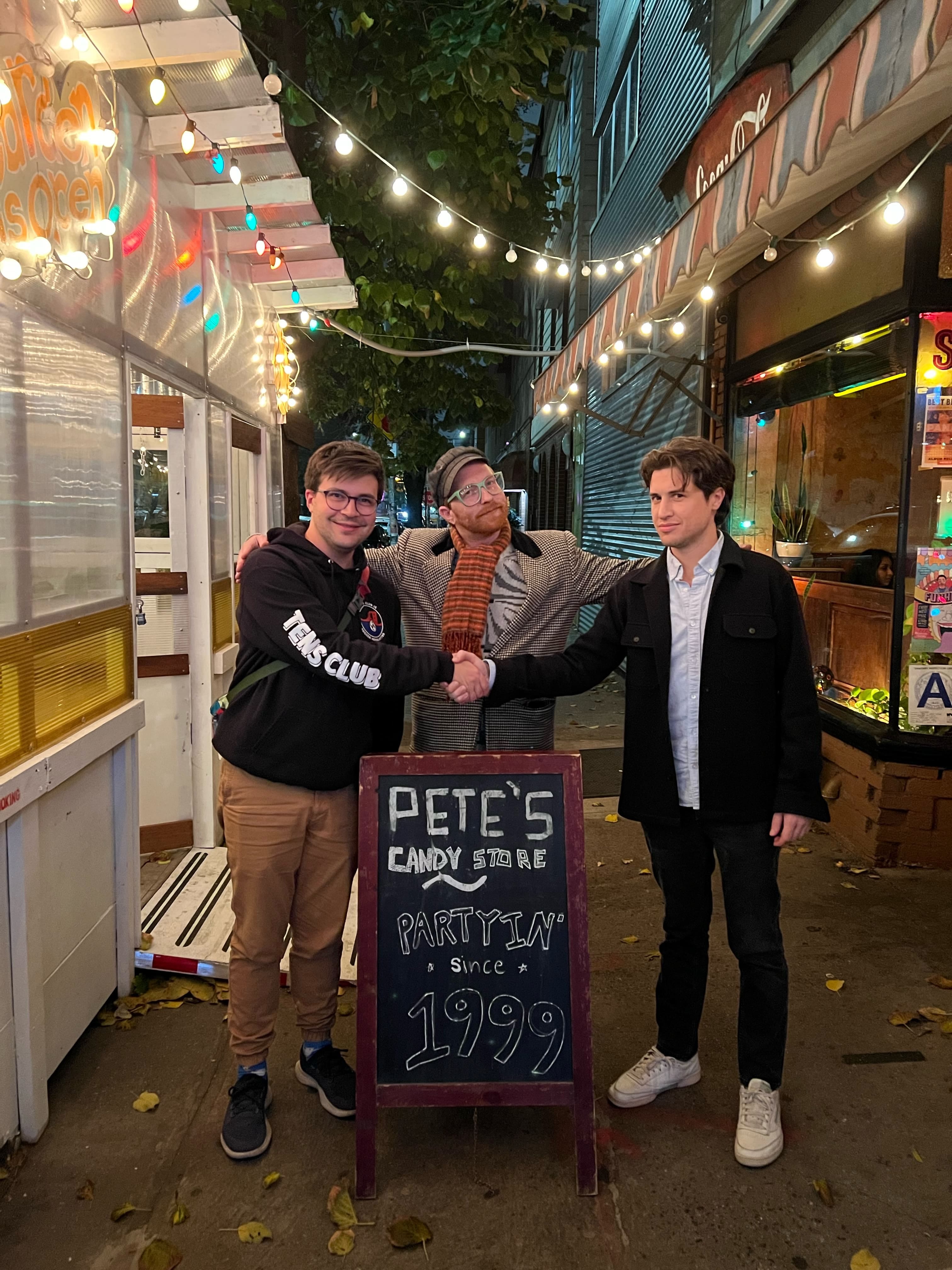 Three white men, all approximately 5'10", stand outside Pete's Candy Store in Greenpoint. The two men on the outer edges shake hands while the third man stands between them with his hands on their shoulders.