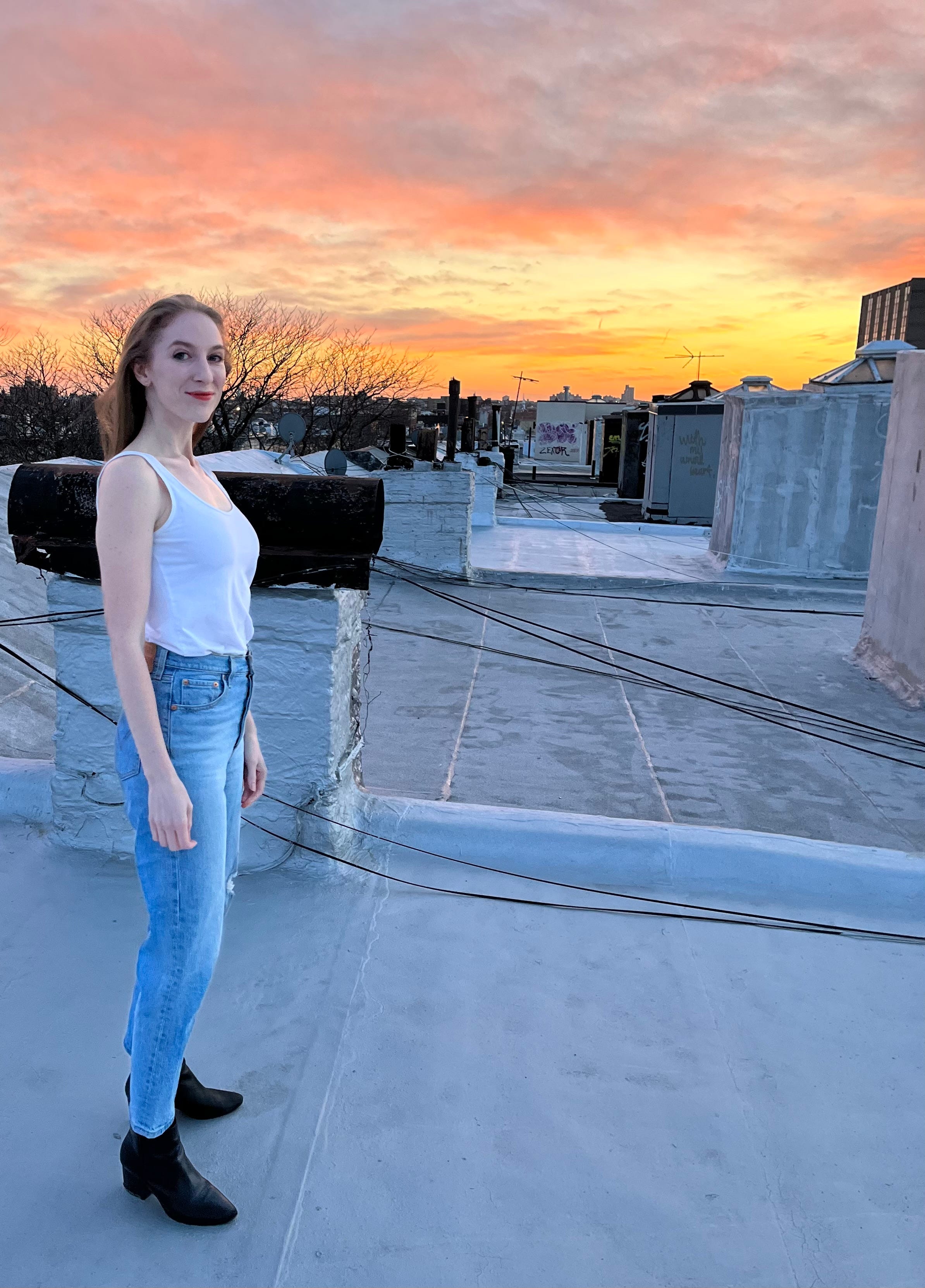 A young woman stands on a rooftop, looking at the camera as the sun sets behind her.