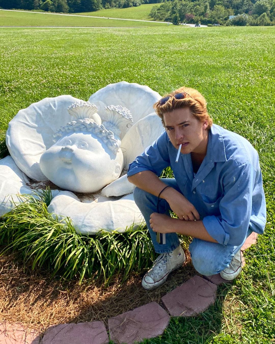 Cole Sprouse, dressed in all denim, kneels beside a stone statue of a Cabbage Patch Kid mid-birth, cigarette perched on the edge of his lips.