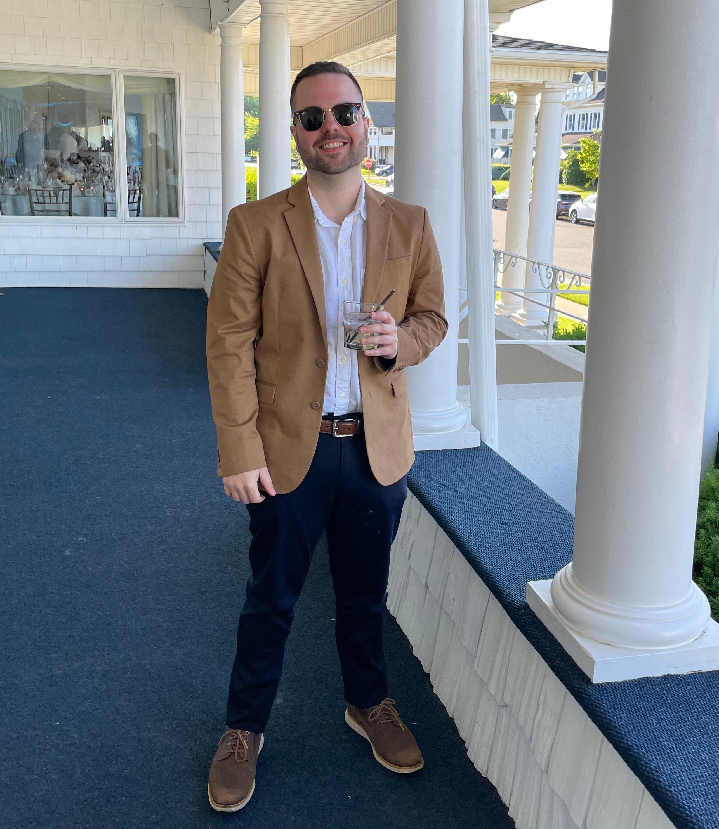 A thirty-something white male in sunglasses and a brown suit jacket stands outside of a wedding holding what appears to be a vodka tonic.
