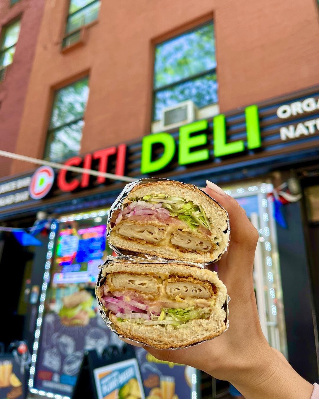 A hand holds up a chicken tender sub, showing off the inside of the sandwich in a cross-section, in front of a bodega.