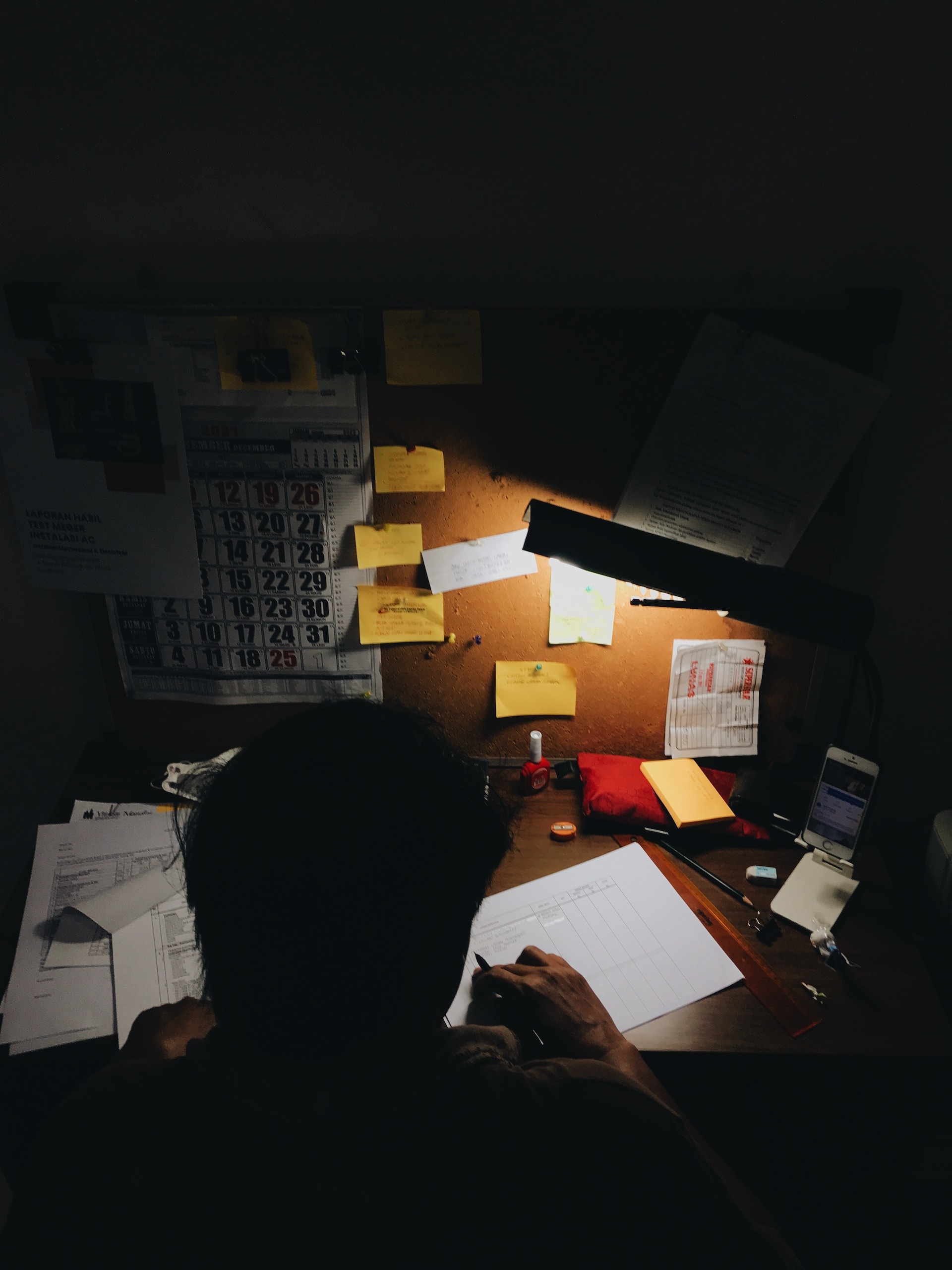 A figure sits at a desk, working in the dark under a lamp.