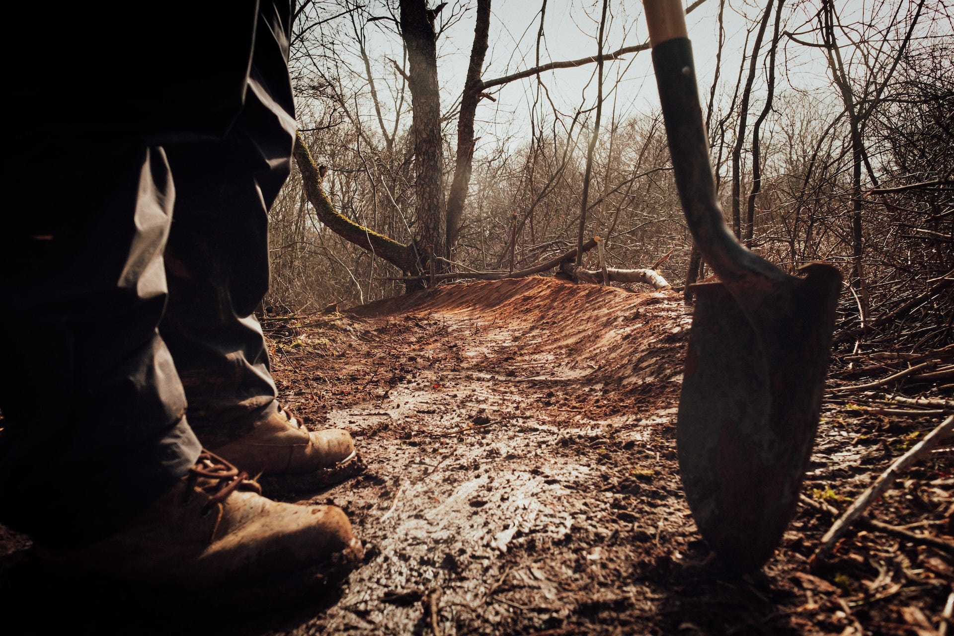 A pair of feet in boots and the tip of a shovel on wooded ground.