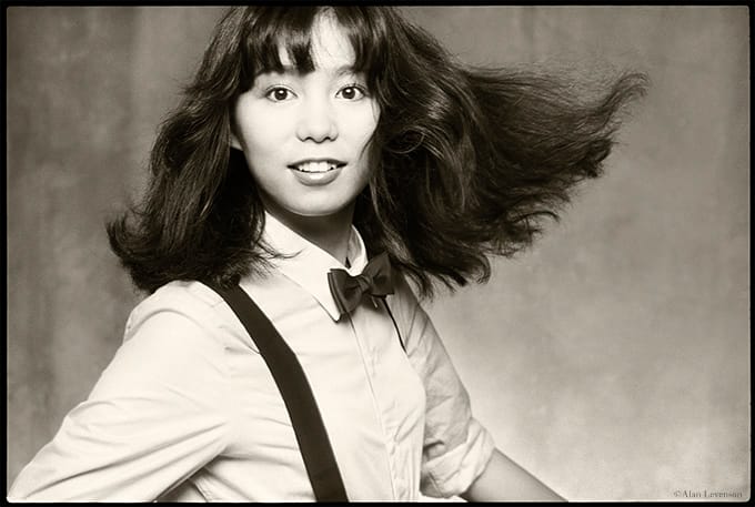 Classic photo of Mariya Takeuchi, in black and white, wearing a bow tie and suspenders, turning towards the camera with her hair flipped out.