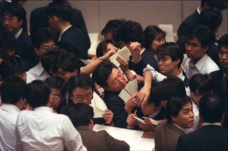 A large group of Japanese business men huddle together with paper and pencil.