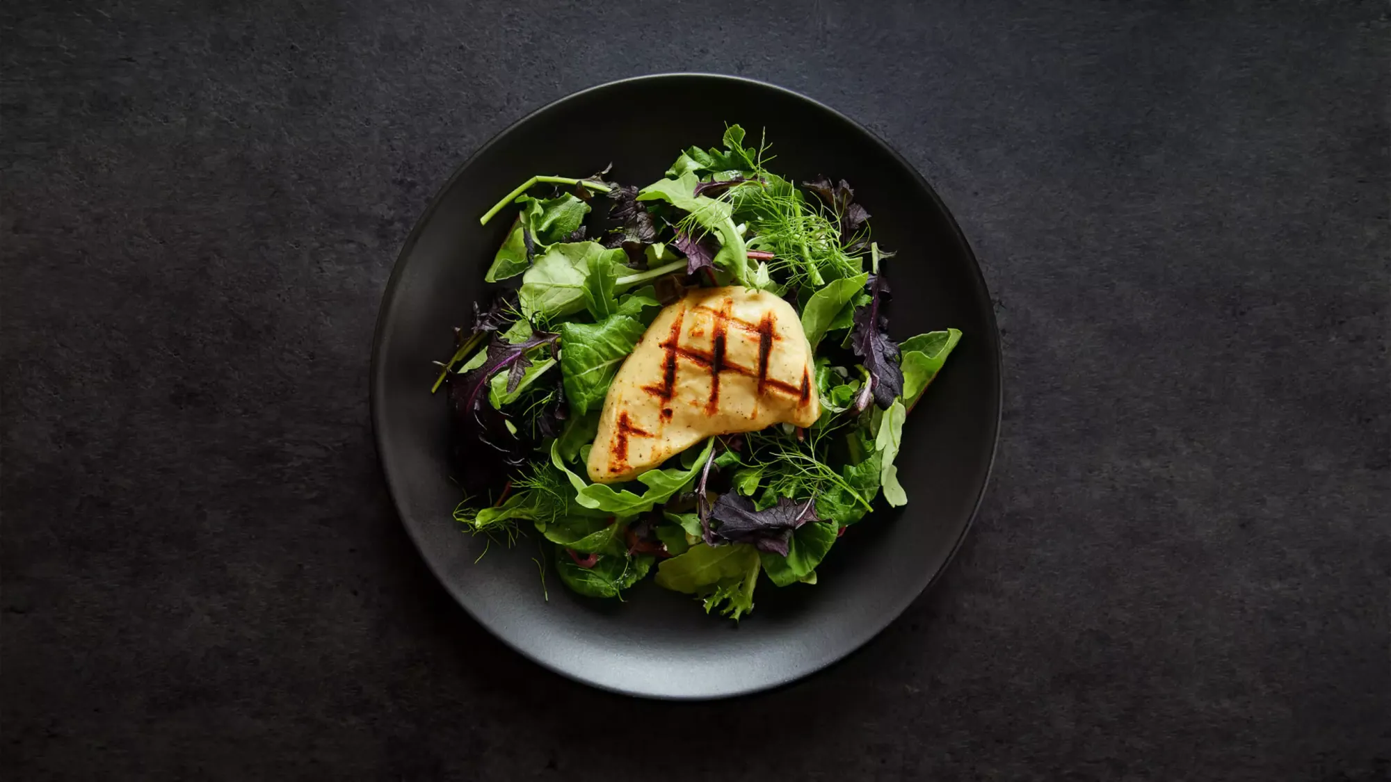 A chicken breast with grill marks sits in the middle of a plate of leafy greens.