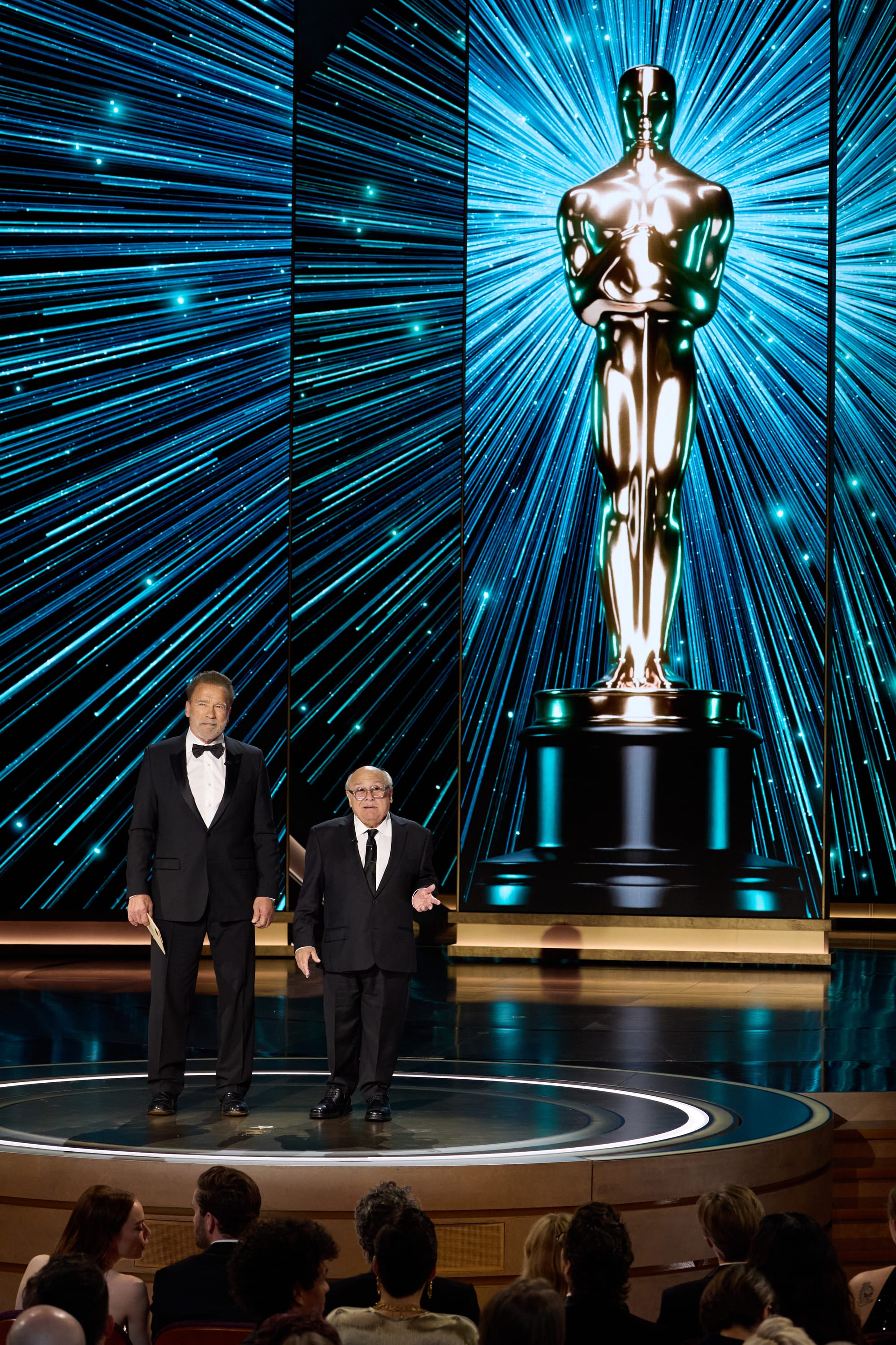 Arnold Schwarzeneger and Danny DeVito stand onstage in front of a large Oscar statuette