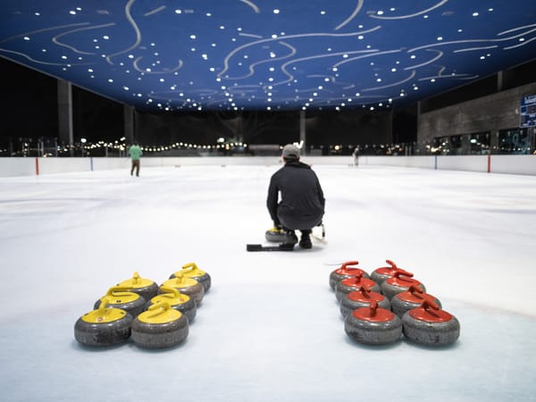 A man in a baseball cap faces away from the camera, preparing to slide a large curling stone down an ice rink.