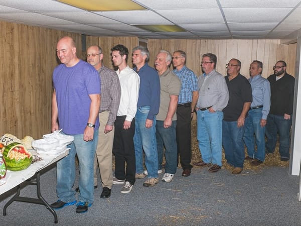 A line of white men of various ages line up at a buffet inside a conference room full of hay.