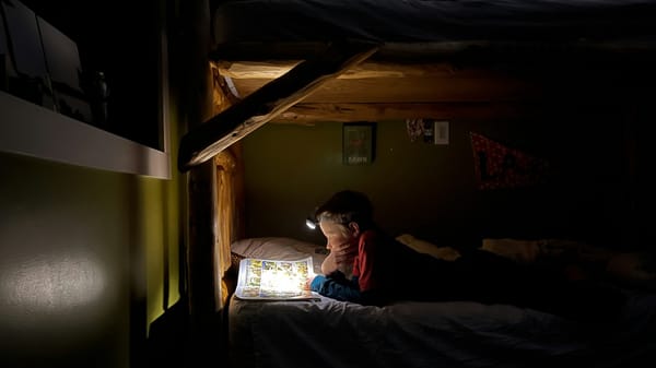 A child reads a comic book in bed with a headlamp.