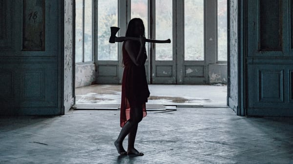 A woman stands barefoot in an abandoned house with an axe.