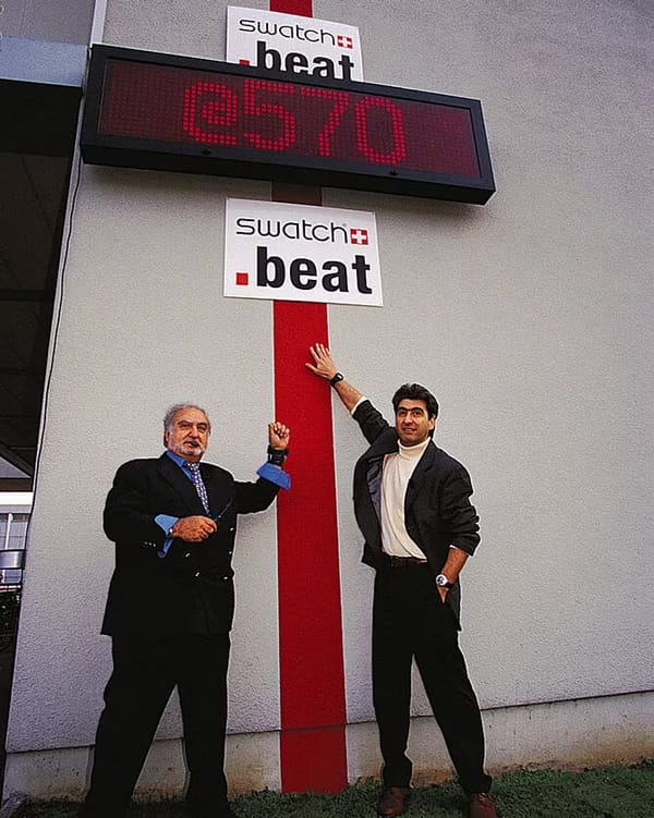 Two men stand in front of a large .beats clock at Swatch HQ.